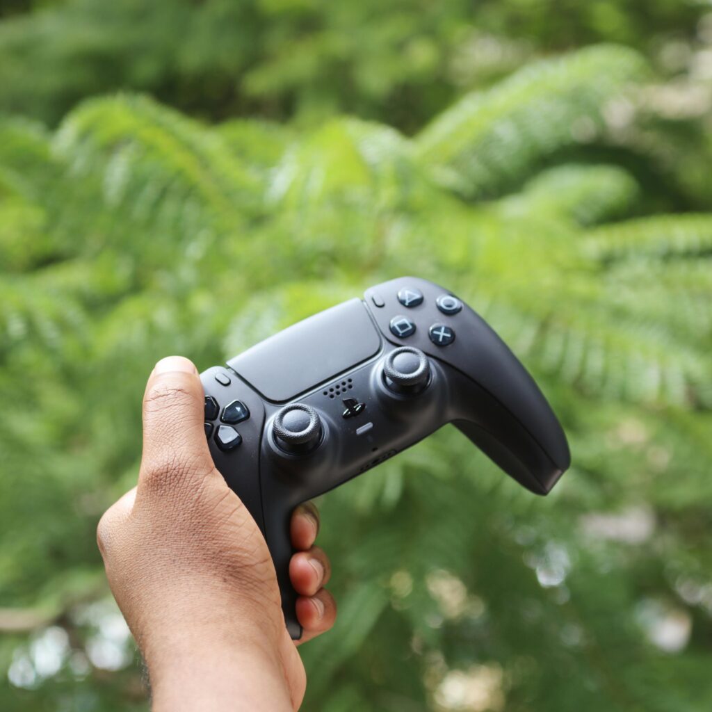 A black wireless game controller being held outdoors against a green leafy background.
